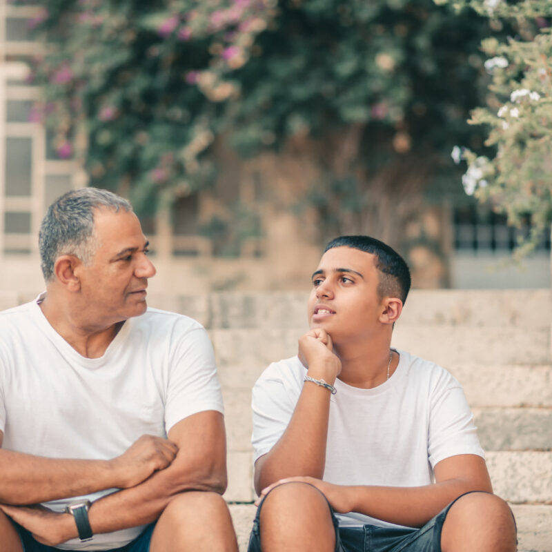 Teenager son and senior father sitting on stairs outdoors at home, talking.